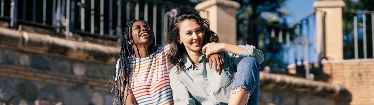 Two friends having fun together on the street sitting on a urban wall. Multiethnic women.