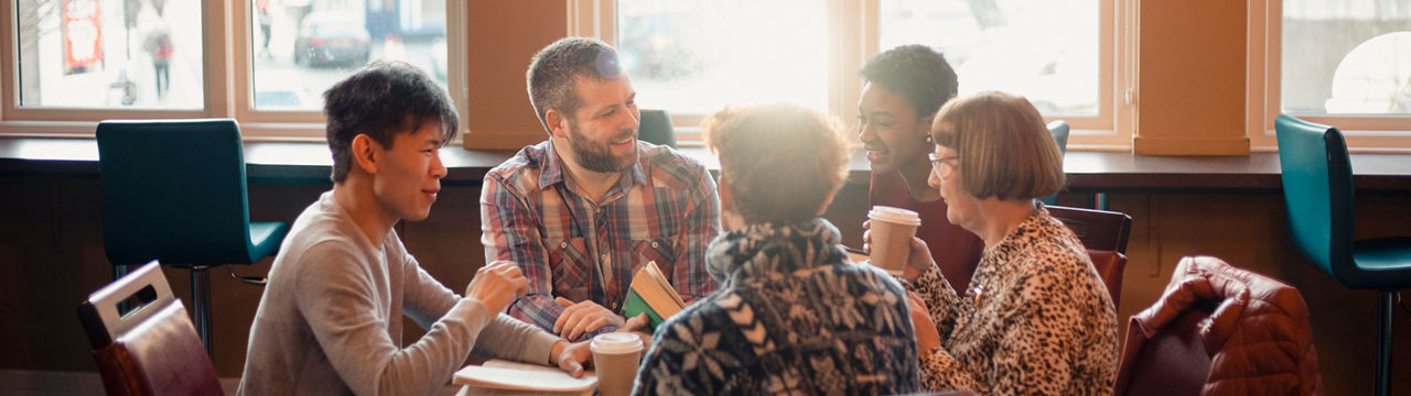 Small group of people with a mixed age range sitting around a table in a cafe. They are talking during a book club.