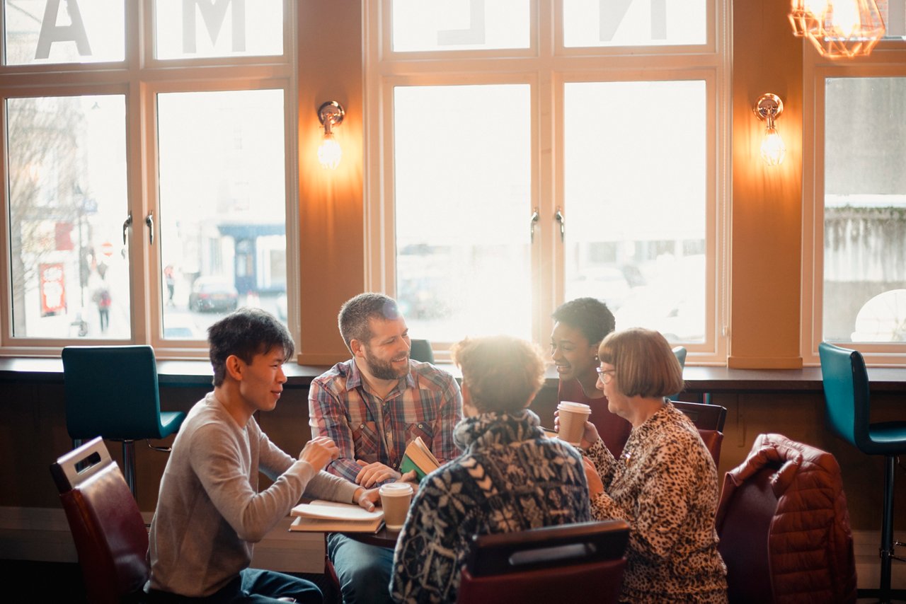 Small group of people with a mixed age range sitting around a table in a cafe. They are talking during a book club.