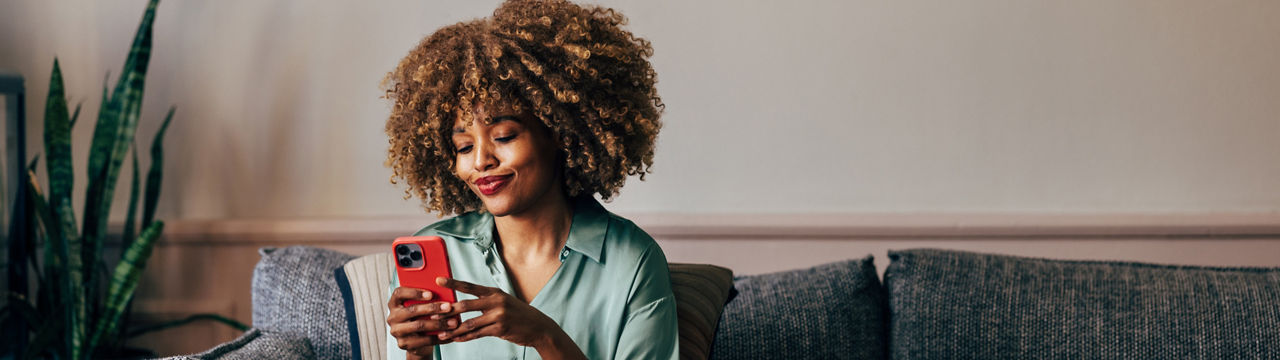 A smiling elegant African-American female using her smartphone while sitting on the cozy sofa in the living room.