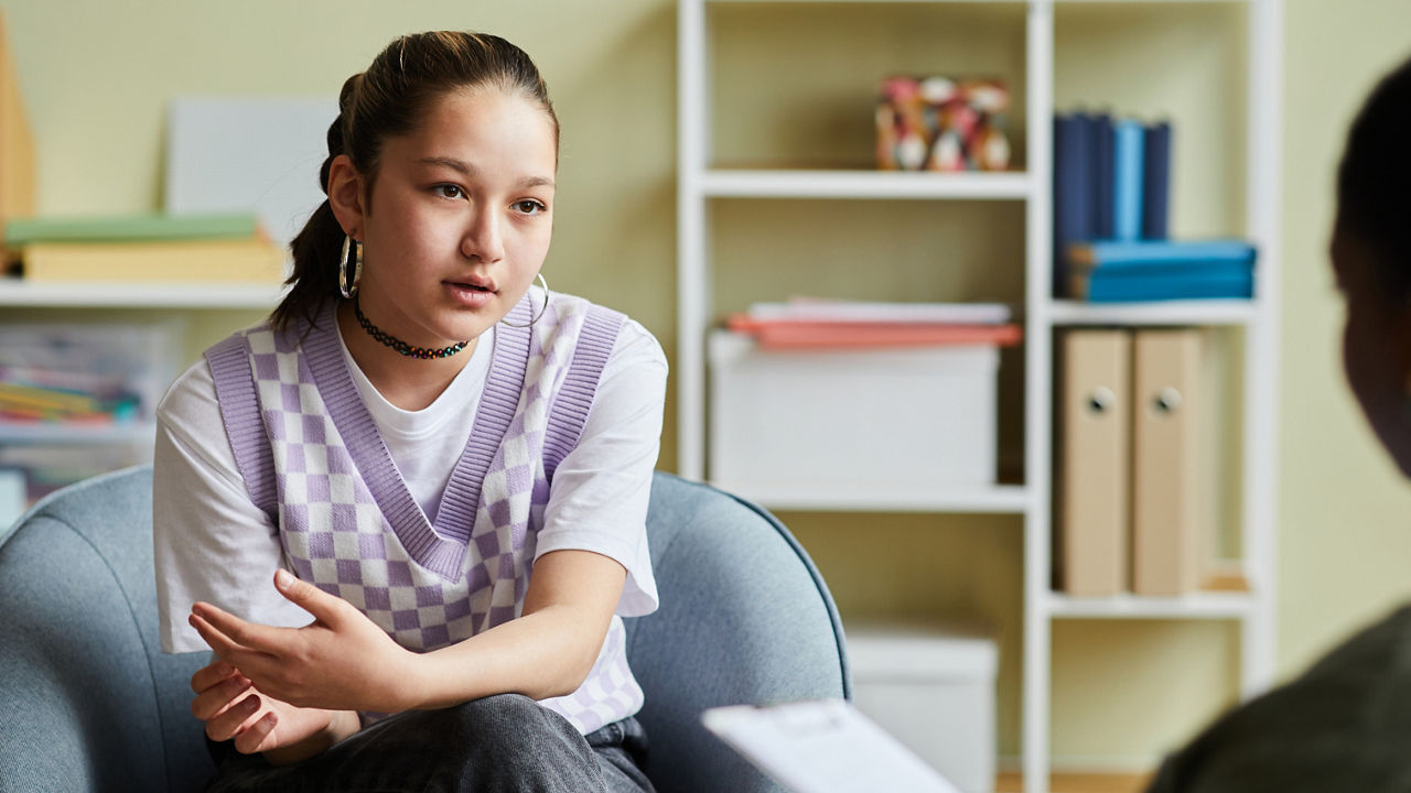 Teenage girl sitting on armchair and talking about her problems to psychologist during consultation at office