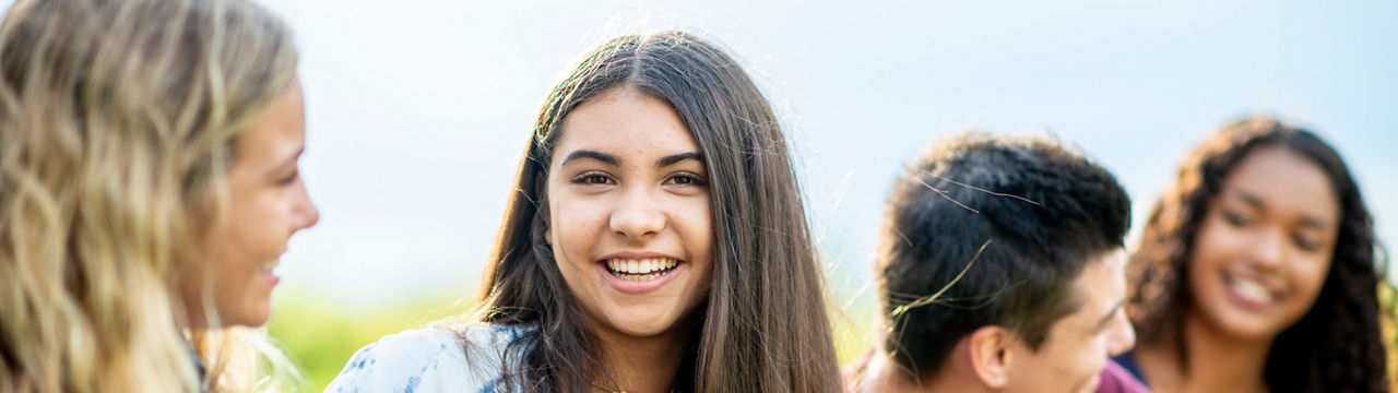 A group of teens are outdoors in a park. They are sitting on the grass, and one girl is smiling at the camera.