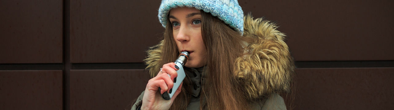 Vape teenager. Young pretty white girl in a cap smoking an electronic cigarette opposite modern brown background on the street in the winter. Bad habit. Close up.