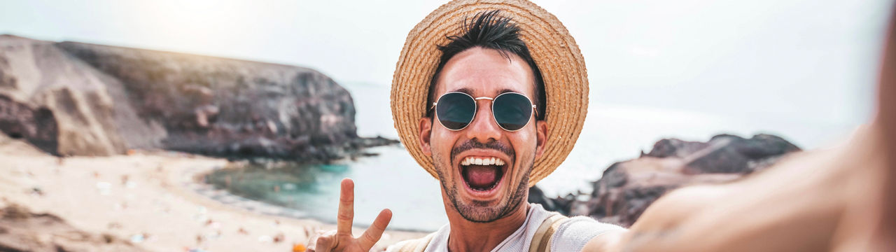 Young man with backpack taking selfie portrait on a mountain - Smiling happy guy enjoying summer holidays at the beach - Millennial showing victory hands symbol to the camera - Youth and journey