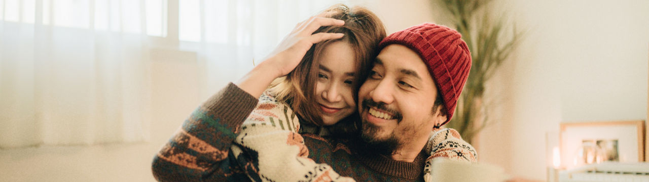 Happy young family couple relaxing talking laughing holding cups drinking coffee tea sitting on bed together in bedroom, loving husband and wife bonding enjoying pleasant conversation at home