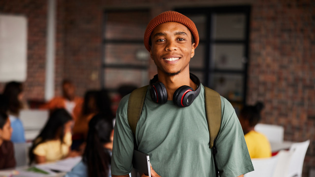 Portrait of a young male college student wearing headphones and a beanie smiling while standing in a classroom with students behind him