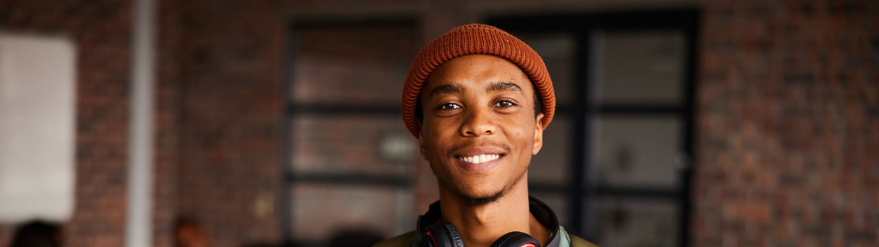 Portrait of a young male college student wearing headphones and a beanie smiling while standing in a classroom with students behind him