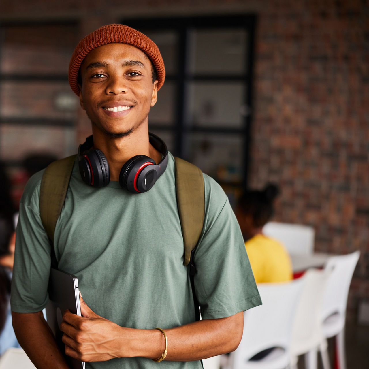 Portrait of a young male college student wearing headphones and a beanie smiling while standing in a classroom with students behind him