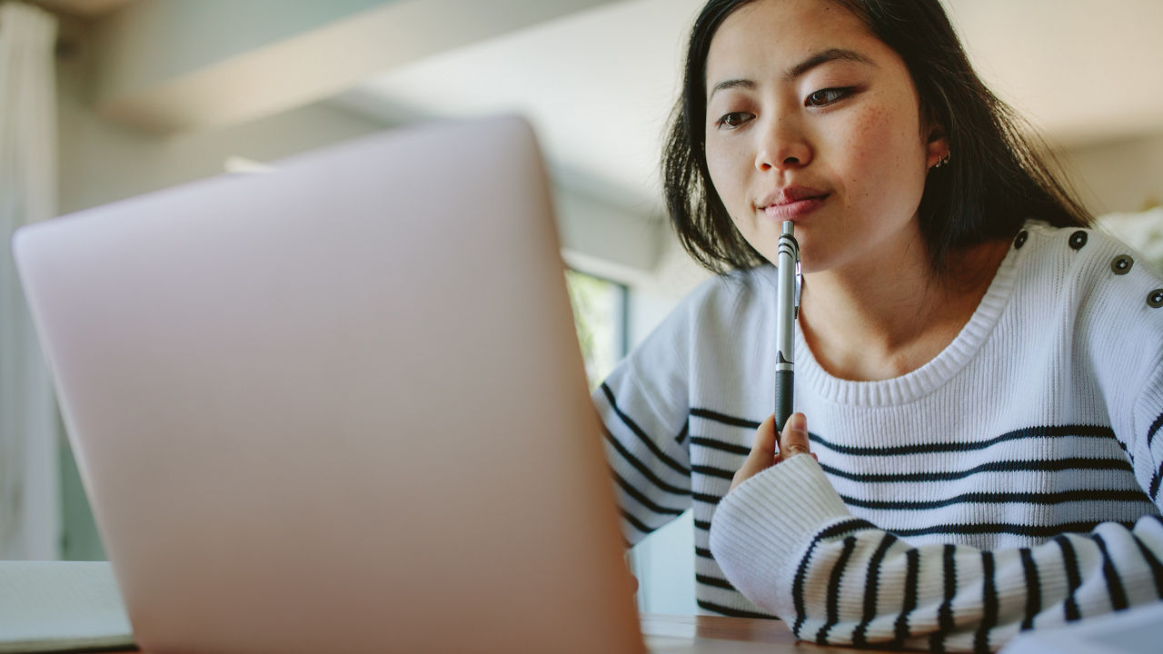 Asian woman studying at home using laptop on the table. Young female using laptop holding a pen in hand