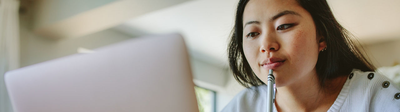 Asian woman studying at home using laptop on the table. Young female using laptop holding a pen in hand