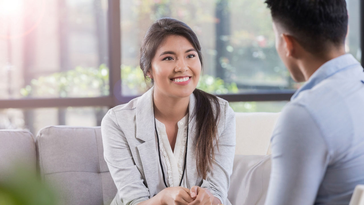 Smiling young Hispanic female therapist attentively listens to male patient discuss something during therapy appointment