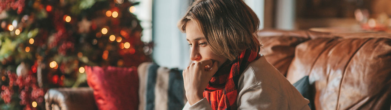 Young teen boy with long hair thoughtful look sad eyes negative mood angry and crying at home. Stylish zoomer gen Z pensive on new year holidays with xmas tree bokeh lights garlands eve 25 december