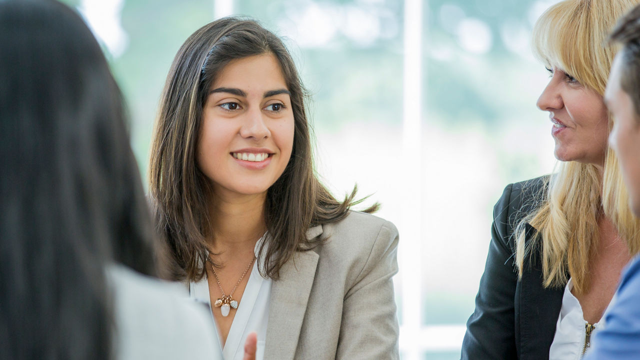 A multi-ethnic group of young business men and women in semi-casual office clothes are sharing ideas in an indoor, sunlit office. A hispanic woman is leading the discussion.
