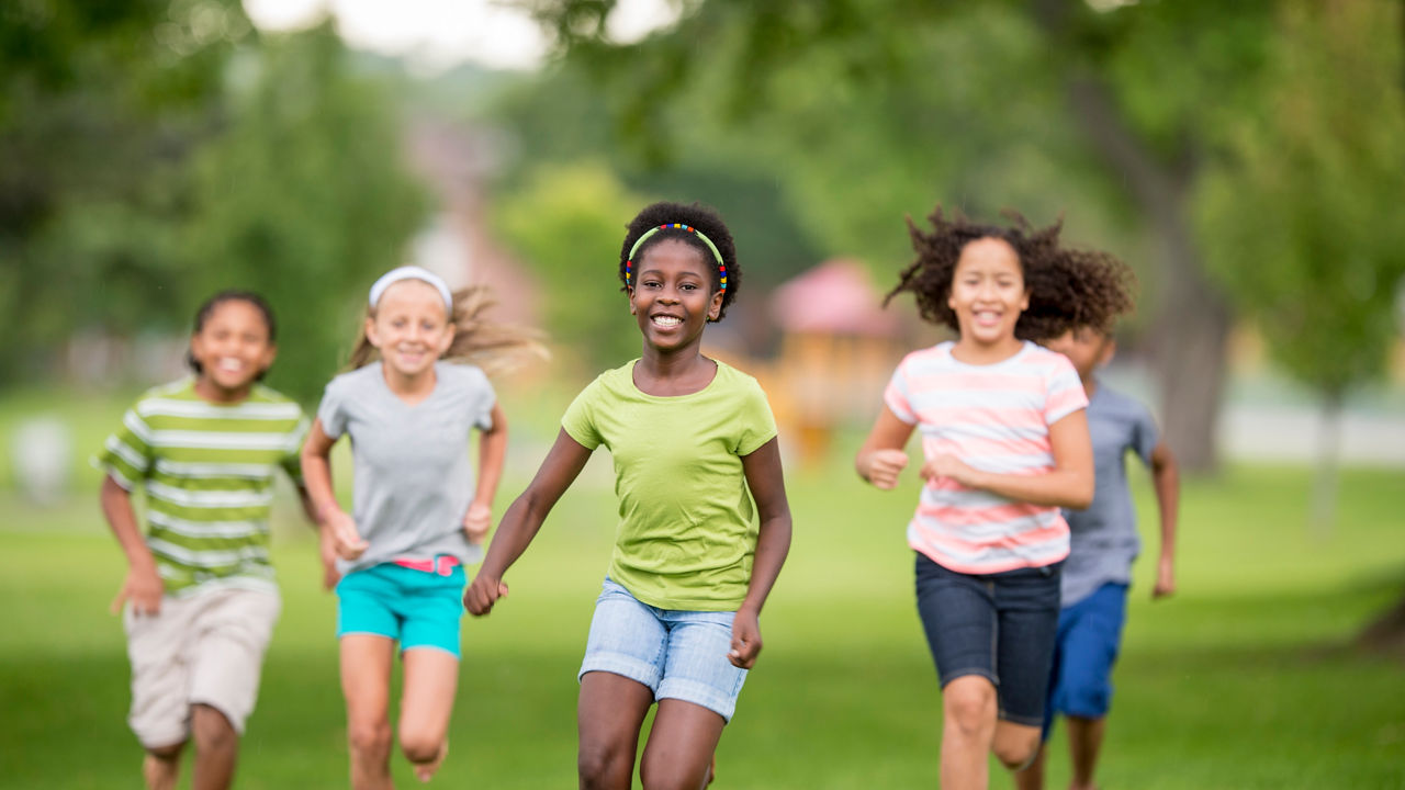 A multi-ethnic group of elementary school children are playing outdoors on a sunny day. They are wearing casual clothing. They are smiling while racing towards the camera.