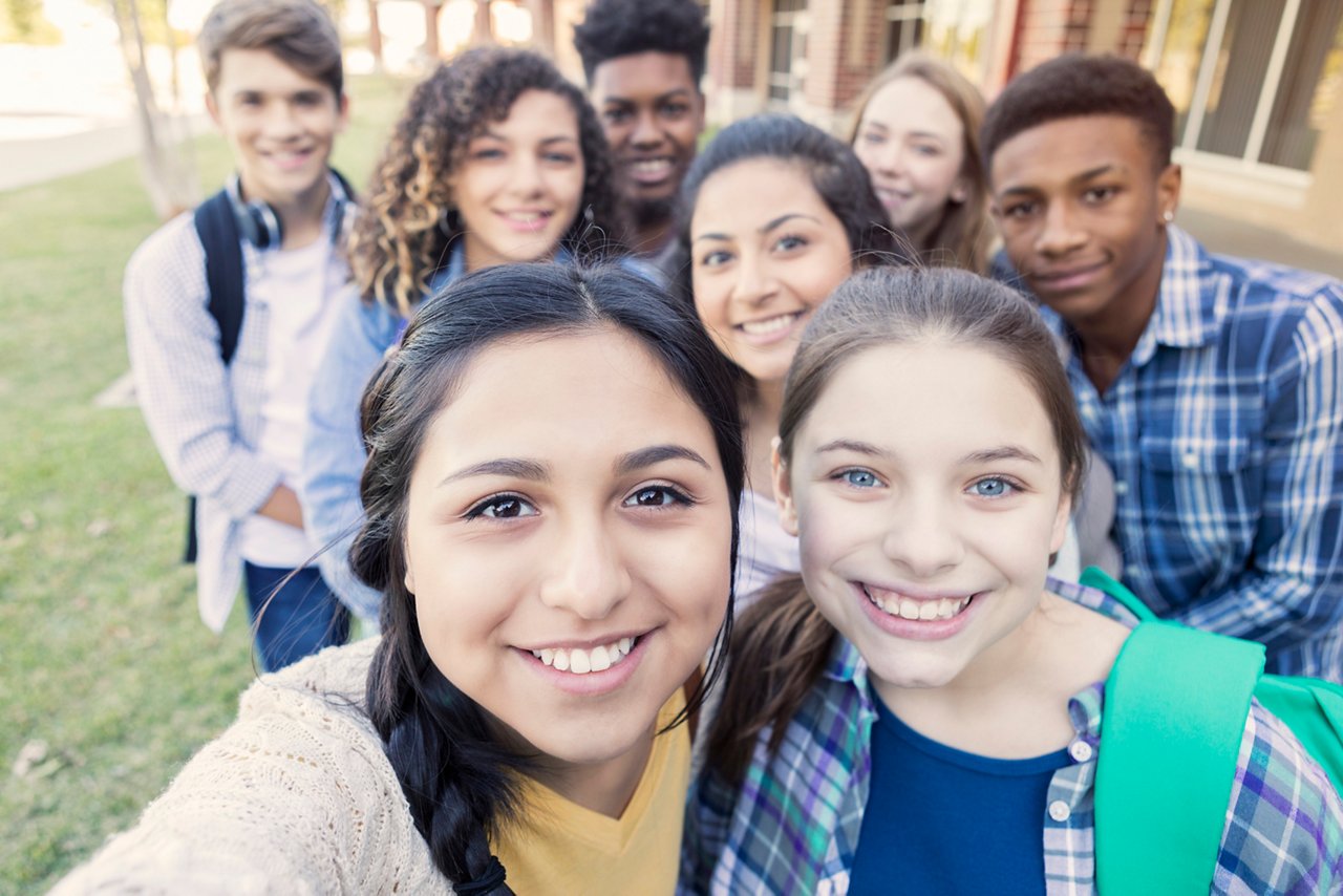 Diverse group of eight high school students are smiling and looking at the camera. Teenagers are students at public high school, and are wearing backpacks or holding school books