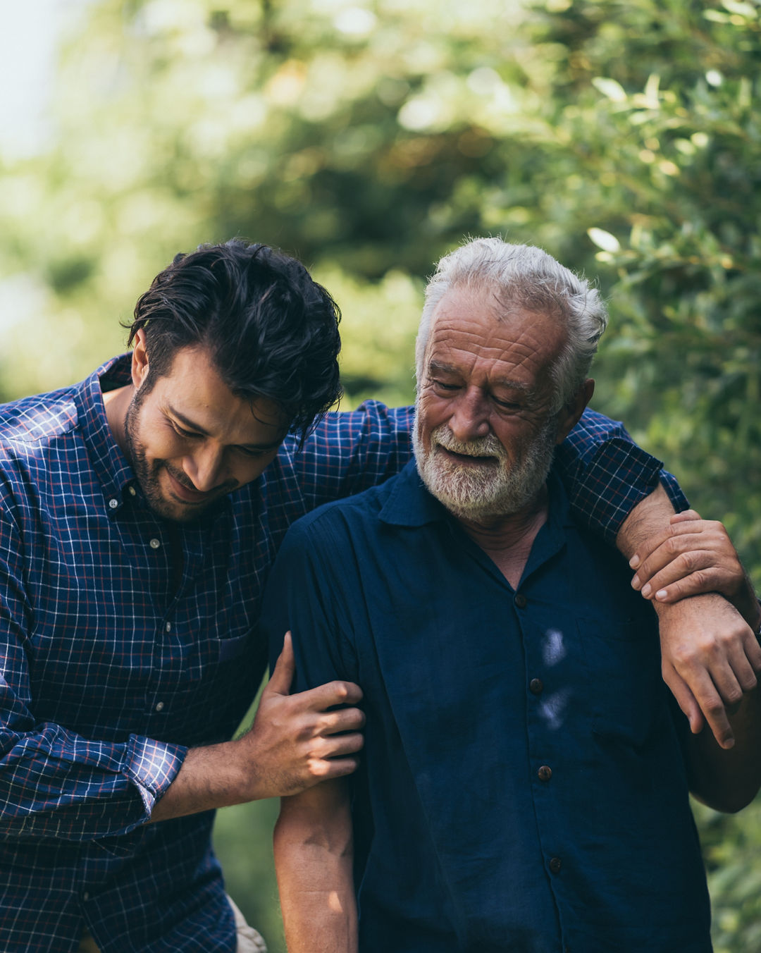 The old man and his son are walking in the park. A man hugs his elderly father. They are happy and smiling.