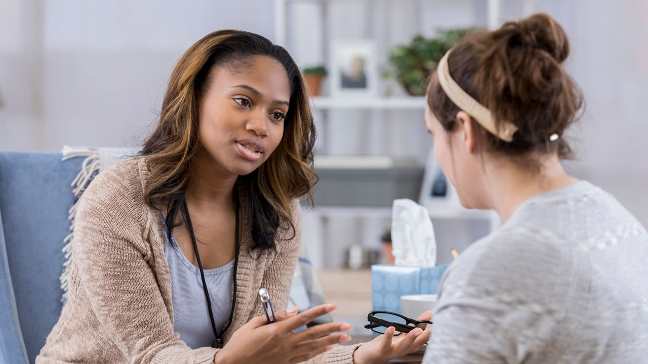 Serious female counselor gestures while talking with Caucasian female client. The counselor is holding eyeglasses and a pen. They are discussing serious issues