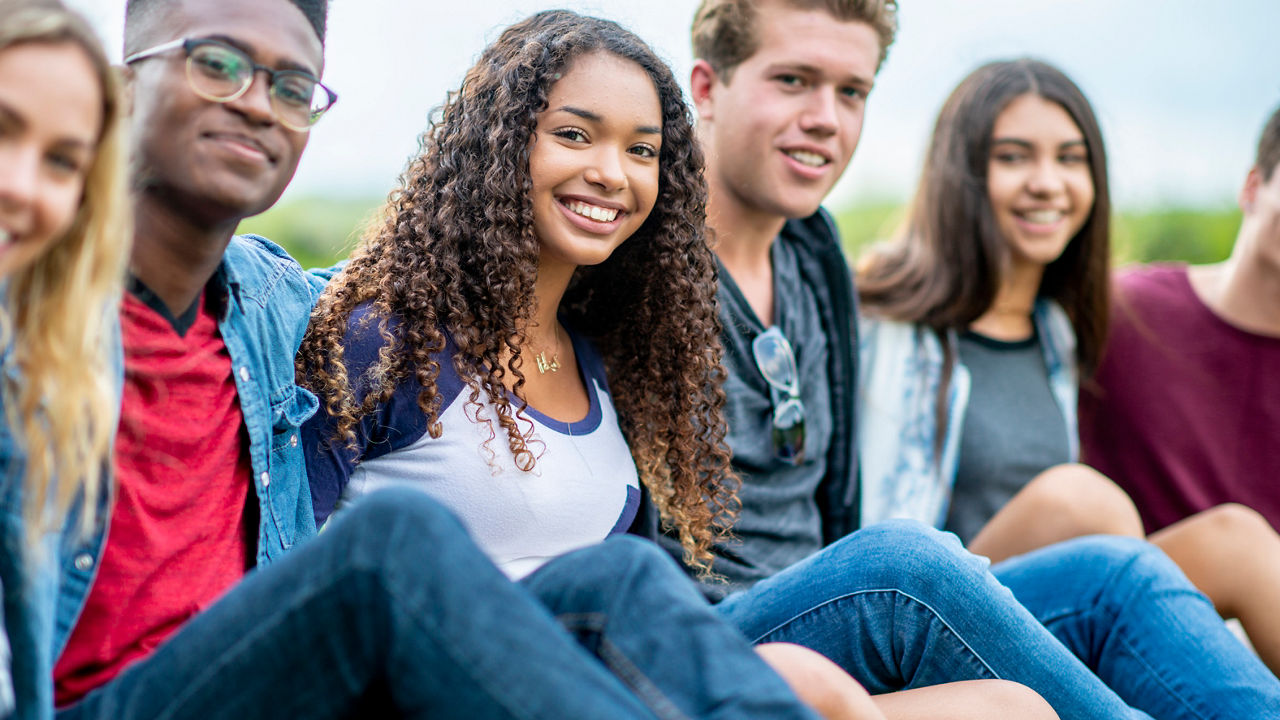 A diverse group of friends sit on the grass with their hands wrapped around each other. They are looking at the camera and smiling, multi-ethnicity, creative content brief