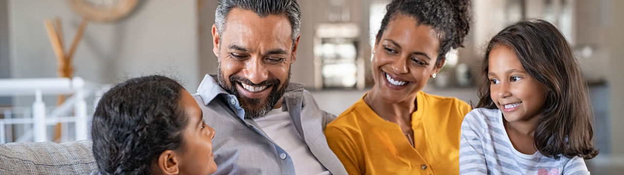 Cheerful multiethnic family at home sitting on sofa listening to daughter. Happy couple with two girls relaxing at home together. Smiling indian parents talking to their daughter at home in the living room