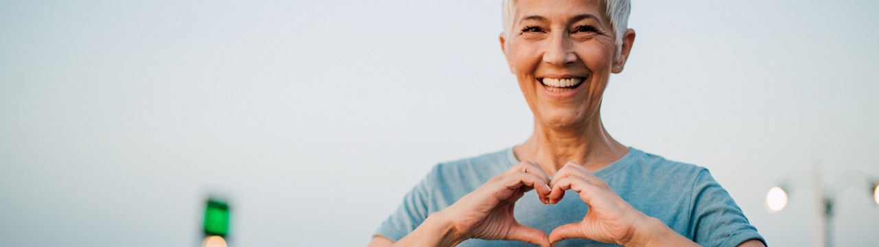 Portrait of a happy senior woman making a heart shape with her hands after exercising on the riverbank