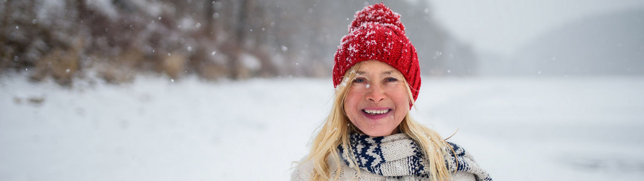 A front view portrait of happy senior woman with hat and outdoors standing in snowy nature, looking at camera