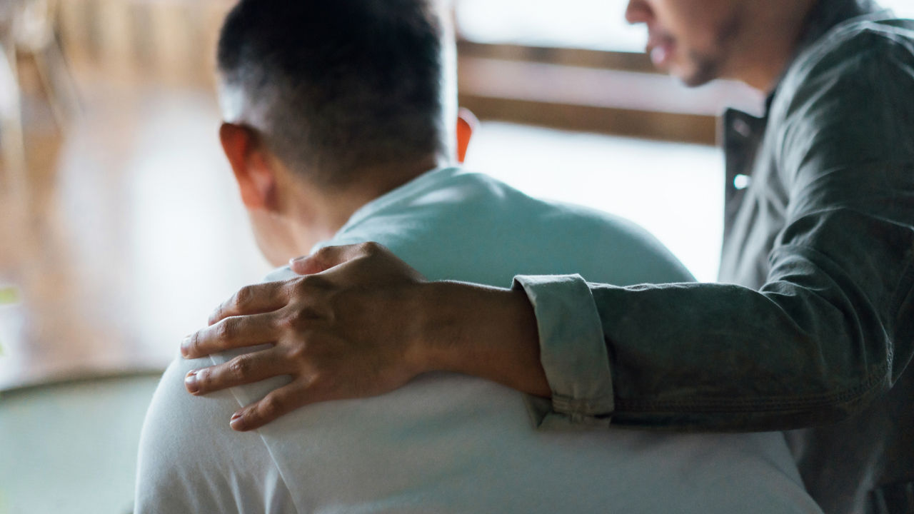 Rear view of son and elderly father sitting together at home. Son caring for his father, putting hand on his shoulder, comforting and consoling him. Family love, bonding, care and confidence