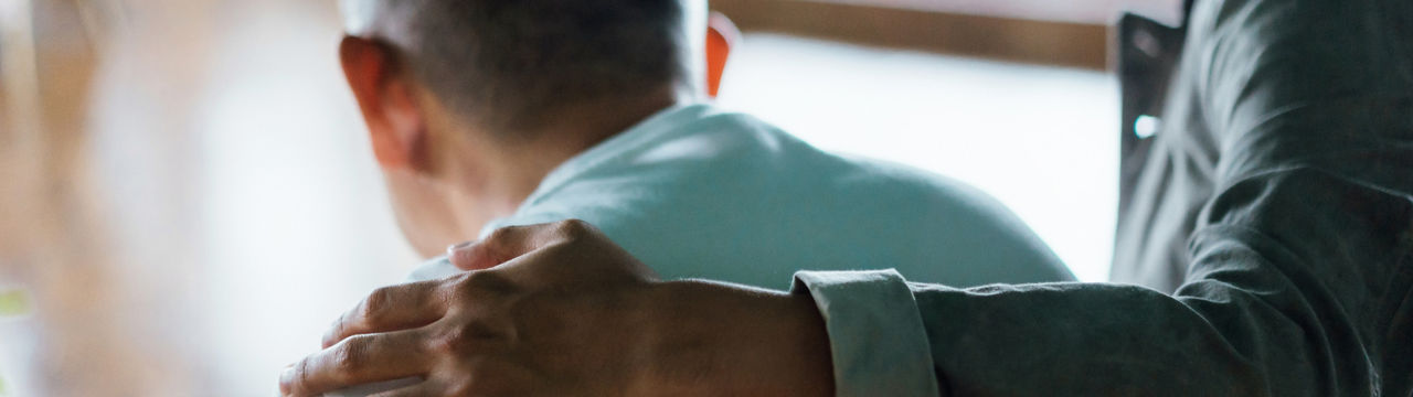 Rear view of son and elderly father sitting together at home. Son caring for his father, putting hand on his shoulder, comforting and consoling him. Family love, bonding, care and confidence