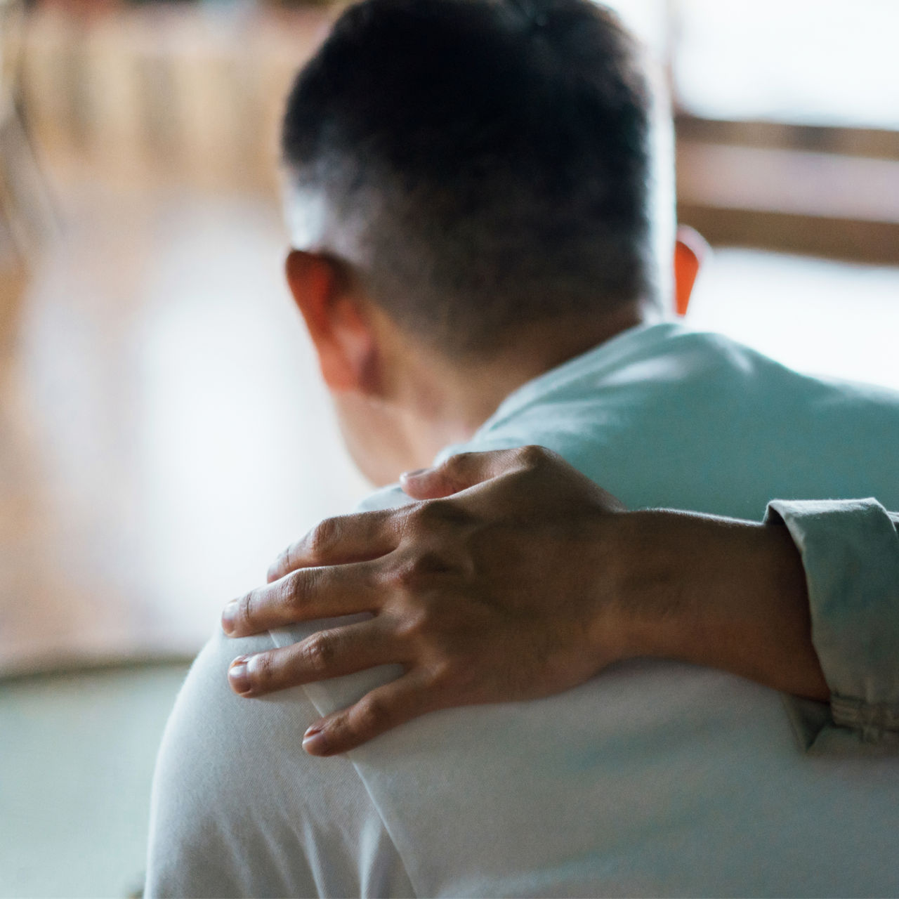 Rear view of son and elderly father sitting together at home. Son caring for his father, putting hand on his shoulder, comforting and consoling him. Family love, bonding, care and confidence