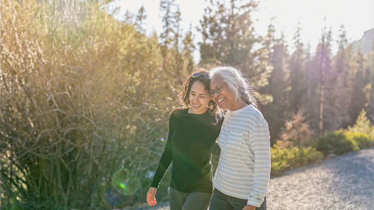 A beautiful mixed race young adult woman embraces her vibrant retirement age mother. The mother and daughter are enjoying a relaxing walk in nature on a beautiful, sunny day. In the background is a mountainous evergreen forest bathed in sunlight