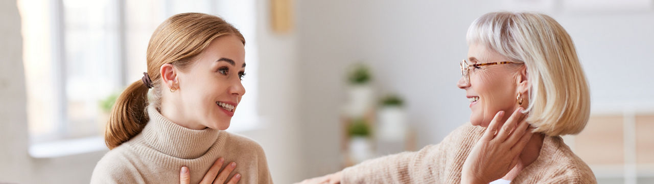 Young woman smiling and talking with senior mother while sitting on comfortable sofa at home together