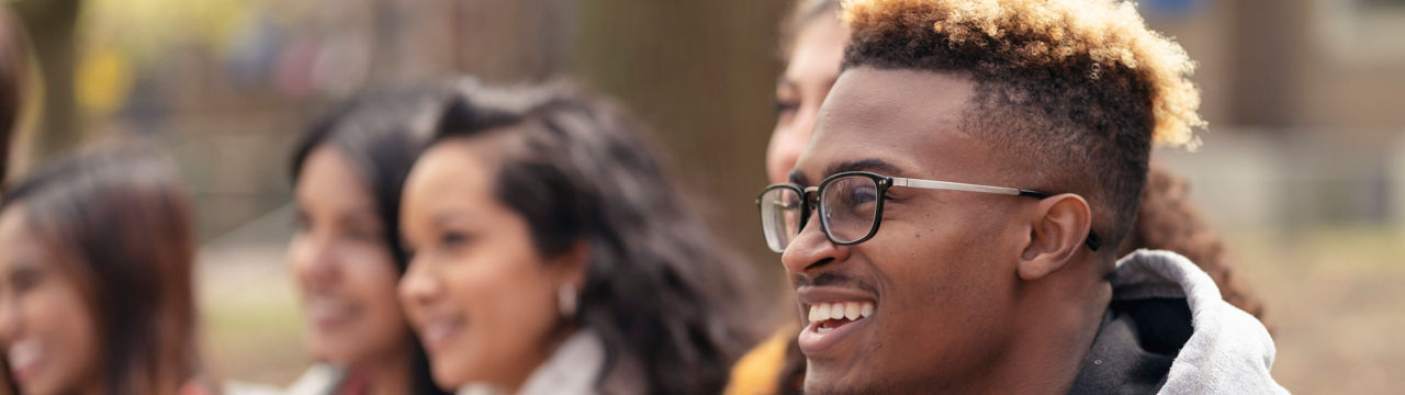 A middle sized group of university aged students, both male and female, are sitting outside on the campus grounds and relaxing before class. They are dressed in casual but stylish clothing while smiling