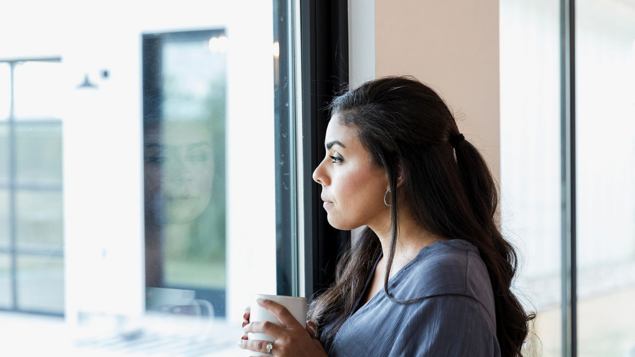 A pensive mid adult woman daydreams as she looks through a window in her home. She is holding a hot beverage.