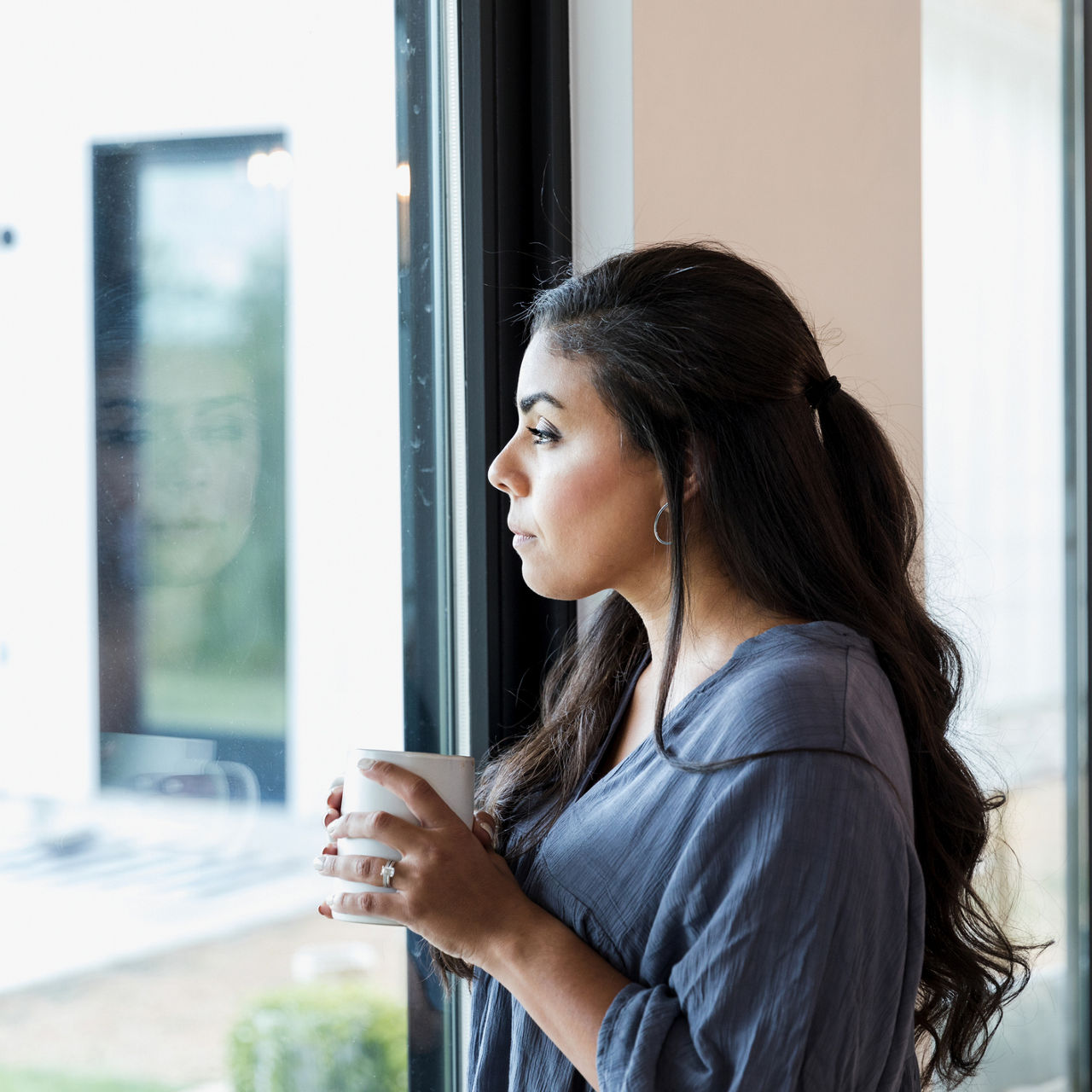 Pensive mid adult woman daydreaming as she looks through a window in her home. She is holding a hot beverage.