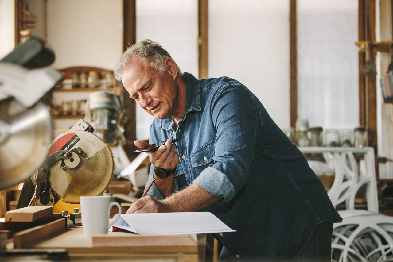 Senior carpenter working at his workshop while standing at desk using mobile phone and writing in a book. Mature man working in carpentry workshop