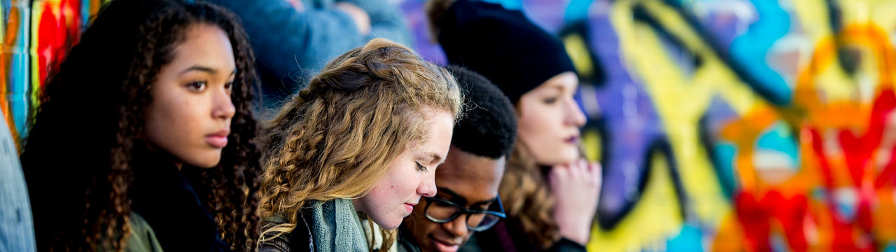 A group of teenagers are sitting in front of a wall covered in graffiti. They are wearing stylish clothes. A boy and girl are looking at a smartphone screen together