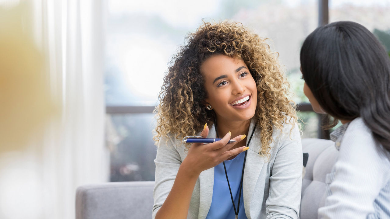 Positive young female therapist gestures as she talks with a female client. The therapist smiles warmly as she talks with the young woman