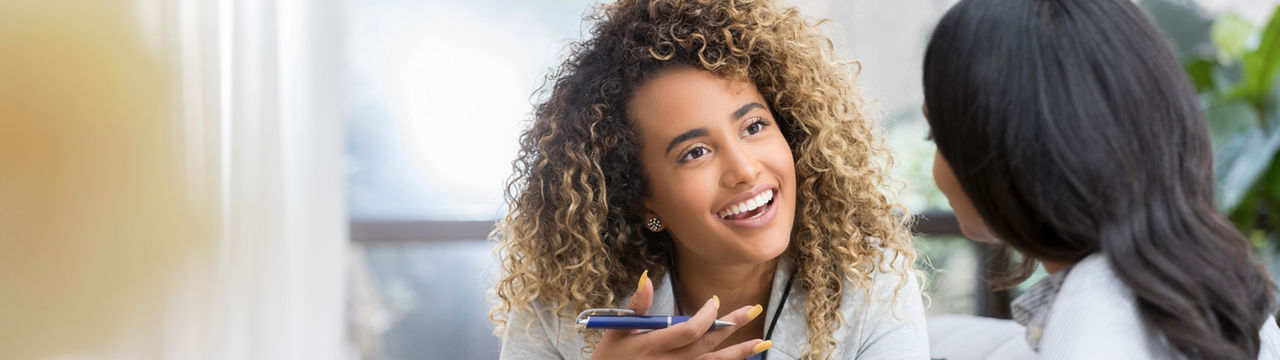 Positive young female therapist gestures as she talks with a female client. The therapist smiles warmly as she talks with the young woman