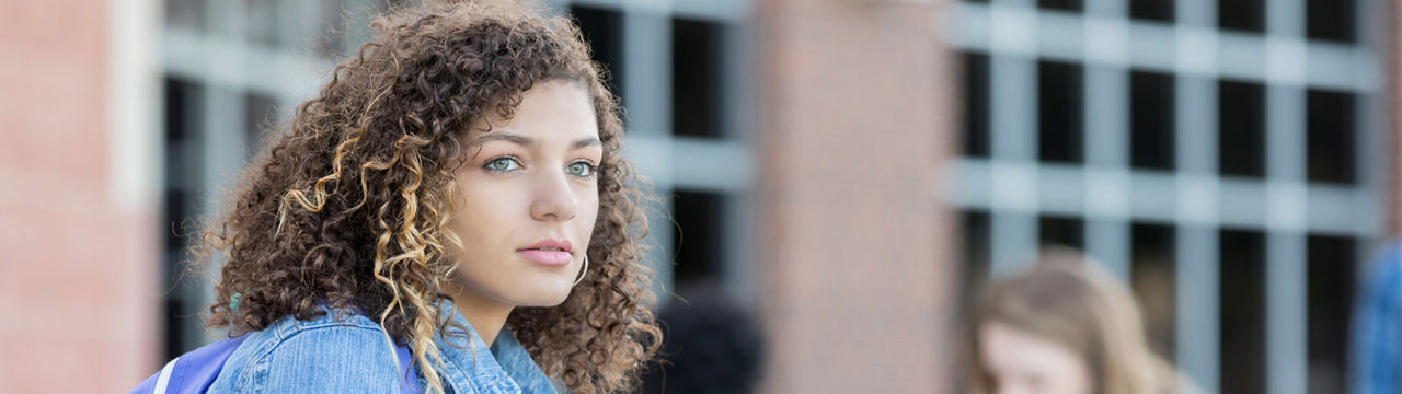 Pensive teenage girl sits outdoors alone while waiting for school to start. She has a concerned expression on her face