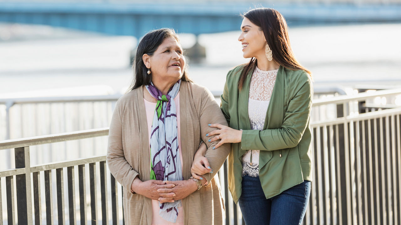 senior Hispanic woman, in her 60s, walking on a city waterfront with her adult daughter, a young woman in her 20s. They are smiling and relaxed, looking at each other as they take a leisurely stroll, extended, Living in Recovery banner