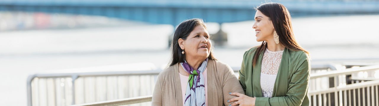 senior Hispanic woman, in her 60s, walking on a city waterfront with her adult daughter, a young woman in her 20s. They are smiling and relaxed, looking at each other as they take a leisurely stroll, extended, Living in Recovery banner