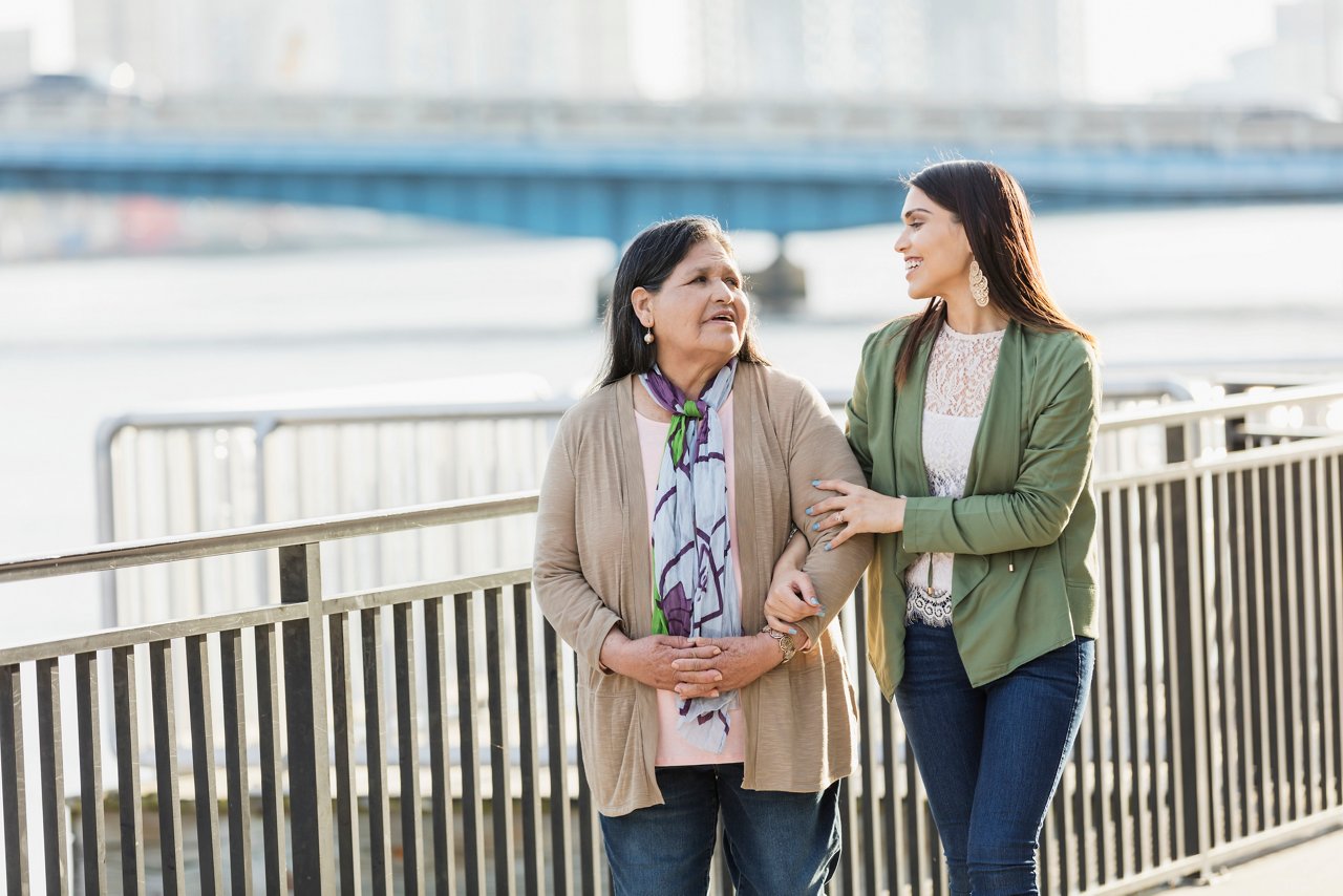 senior Hispanic woman, in her 60s, walking on a city waterfront with her adult daughter, a young woman in her 20s. They are smiling and relaxed, looking at each other as they take a leisurely stroll