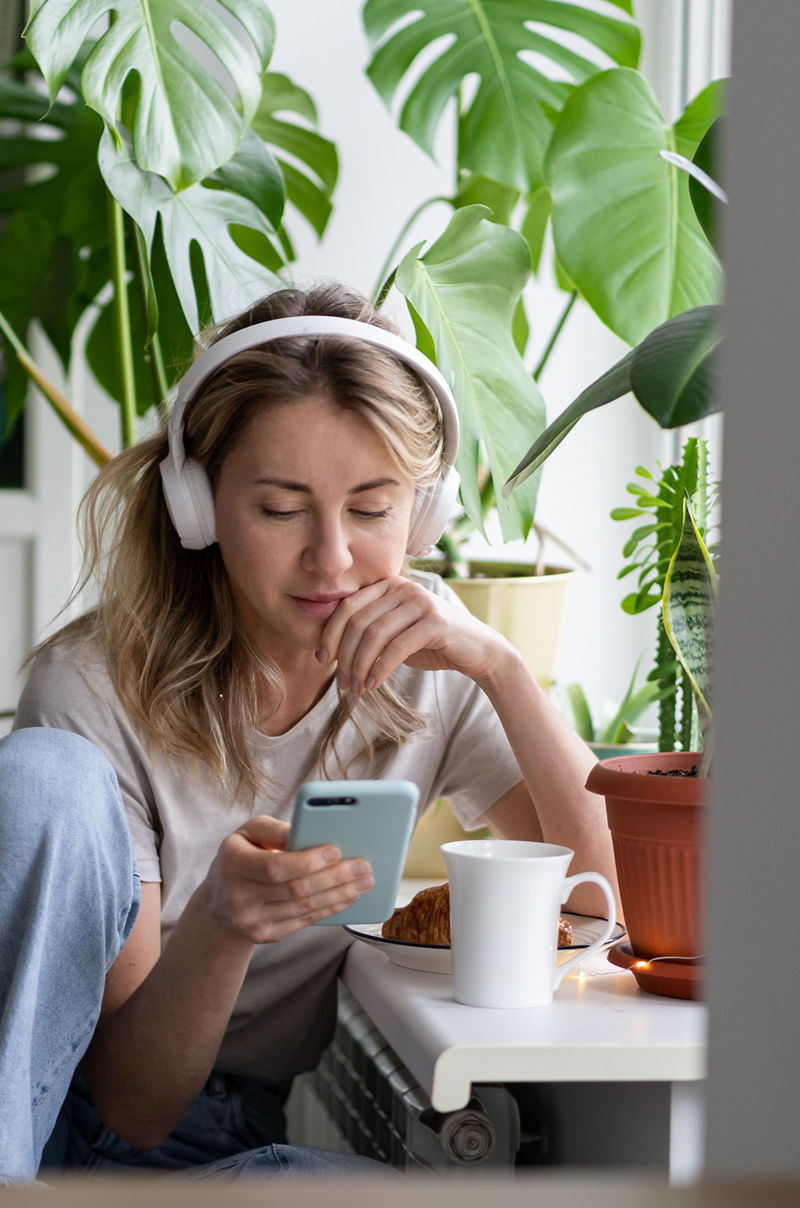 Woman listening to music, wear wireless white headphones, using mobile smart phone, chatting in social networks, sitting next to the window, houseplants on windowsill. Life at home. Time to relax