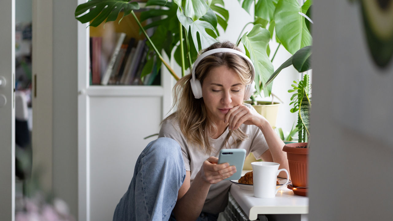 Woman listening to music, wear wireless white headphones, using mobile smart phone, chatting in social networks, sitting next to the window, houseplants on windowsill. Life at home. Time to relax