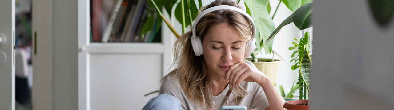 Woman listening to music, wear wireless white headphones, using mobile smart phone, chatting in social networks, sitting next to the window, houseplants on windowsill. Life at home. Time to relax