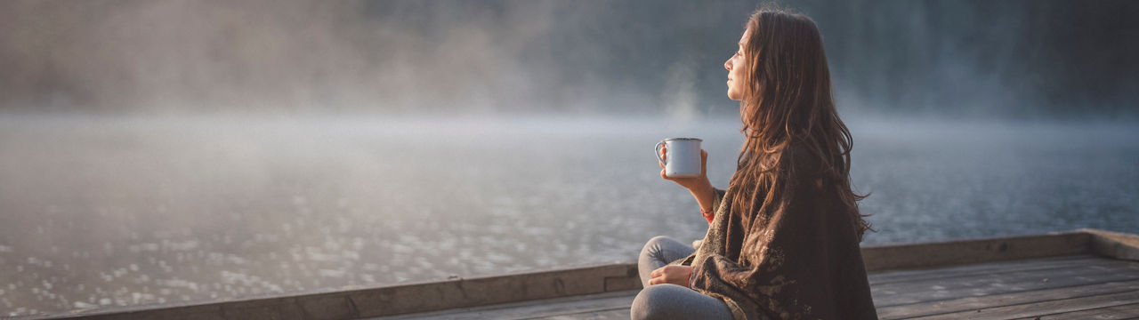 Side view of woman sitting on wooden pier in nature, she looking the sunrise and drinking coffee. 