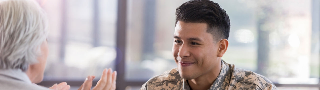 Young adult Hispanic male smiles softy as his female counselor sitting across the table from him. She gestures as she speaks