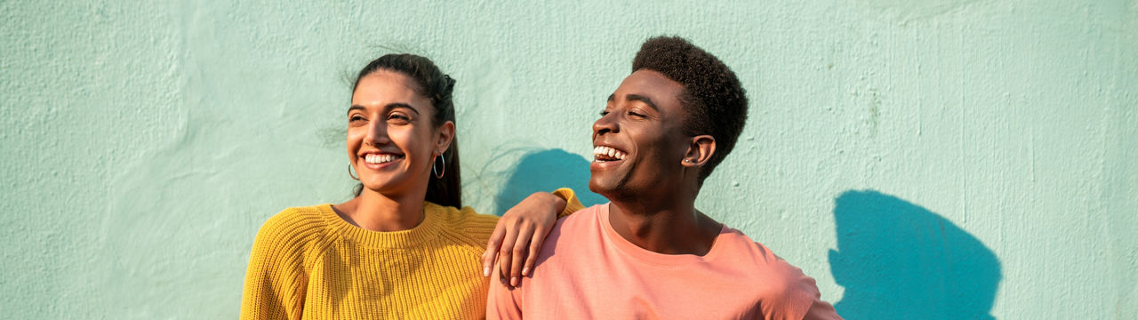 Portrait of young couple. They are looking away laughing and leaning on light blue wall