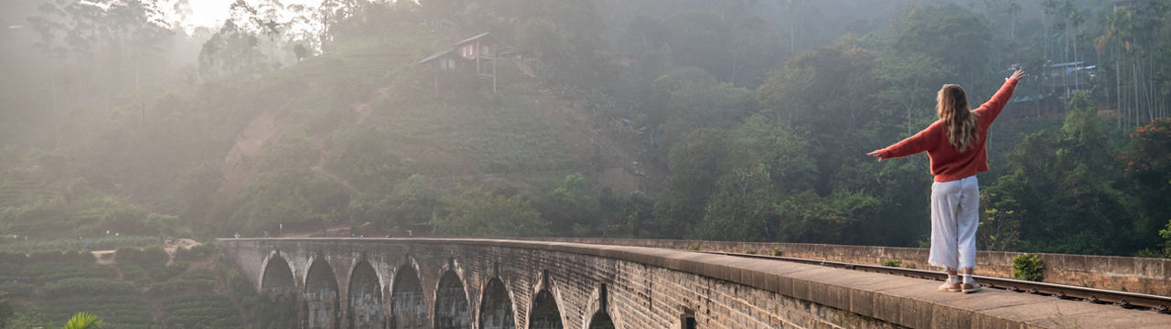 Playful young woman walking on the edge of ancient viaduct bridge at sunrise and balancing with arms outstretched by her side. Girl travel fun Sri Lanka