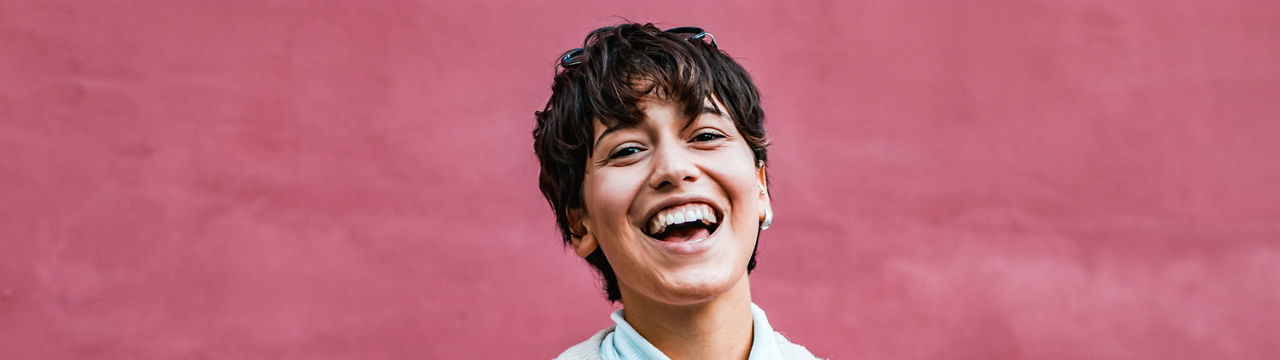 Young girl with short hair standing by the wall while holding a cellphone in her hand and smiling