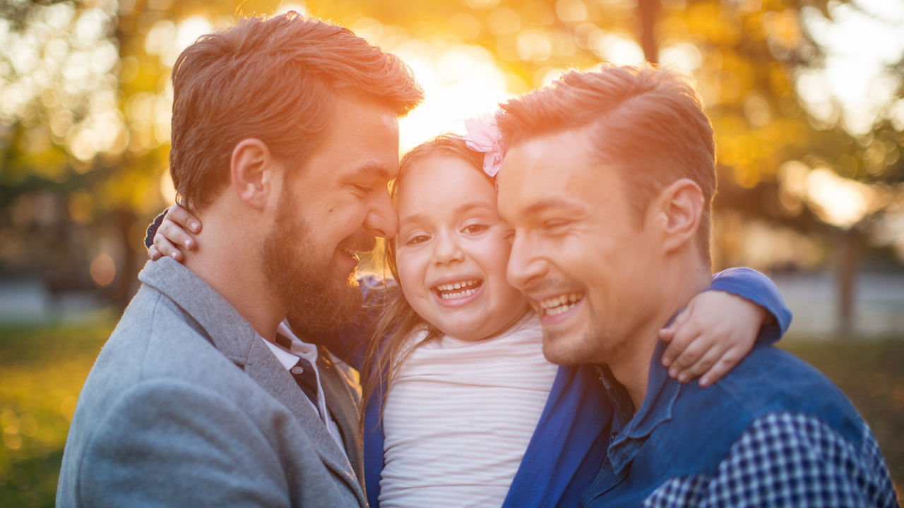 Young gay parents with their daughter having fun in park. Parents holding girl in arms. Enjoying in beautiful sunset.